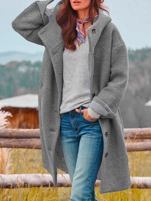 Langer Kapuzenmantel in Grau, offen getragen über grauem Pullover und Jeans, dazu ein buntes Halstuch, Hand an der Kapuze. 2