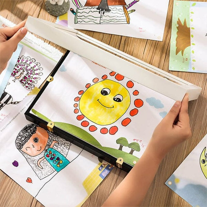 Zwei Hände öffnen einen Rahmen mit Sonnen-Zeichnung auf einem Holztisch, umgeben von weiteren Kinderbildern. 12