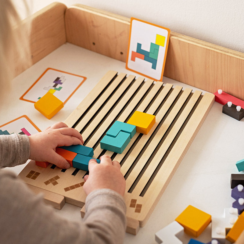 Kinderhände legen farbige Holzteile auf das flache Schlitzbrett, im Hintergrund stehen Vorlagekarten an einer Holzwand. 3