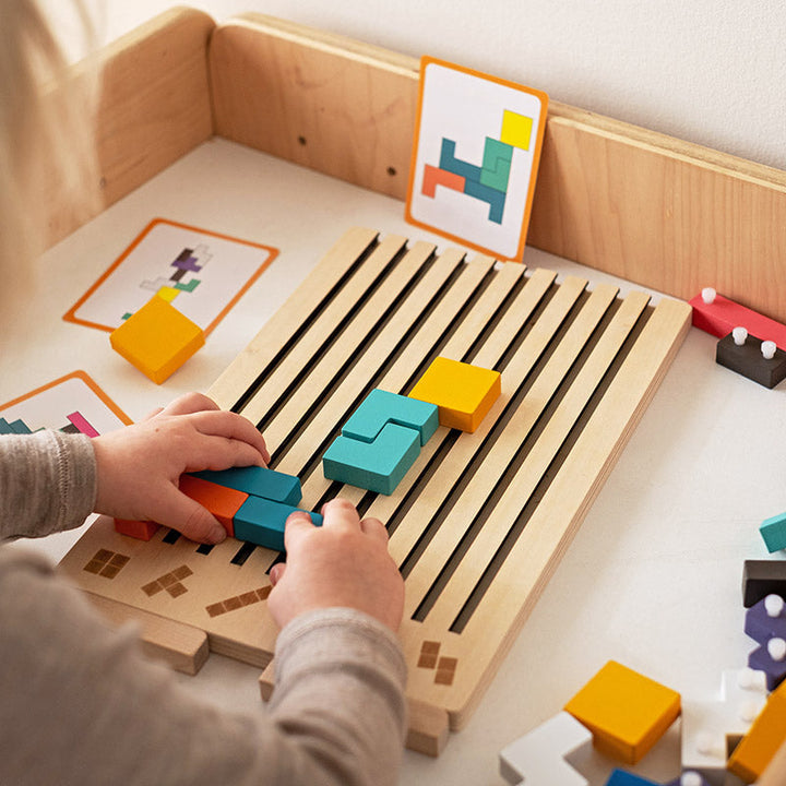Kinderhände legen farbige Holzteile auf das flache Schlitzbrett, im Hintergrund stehen Vorlagekarten an einer Holzwand. 3