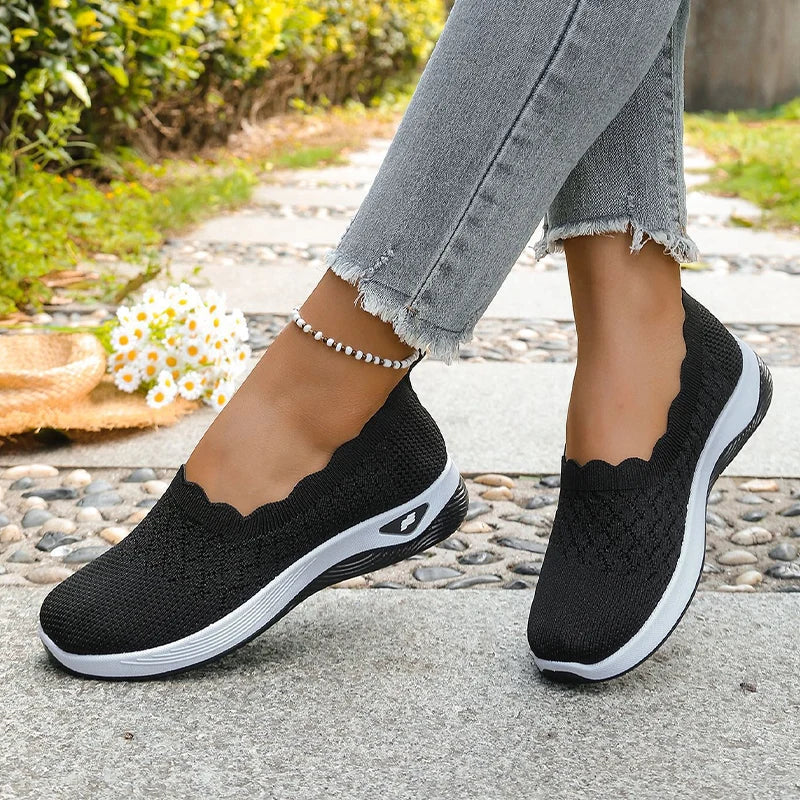 Schwarze Slip-ons mit weißer Sohle, getragen auf einem Weg mit Kieselsteinen und Blumen 14