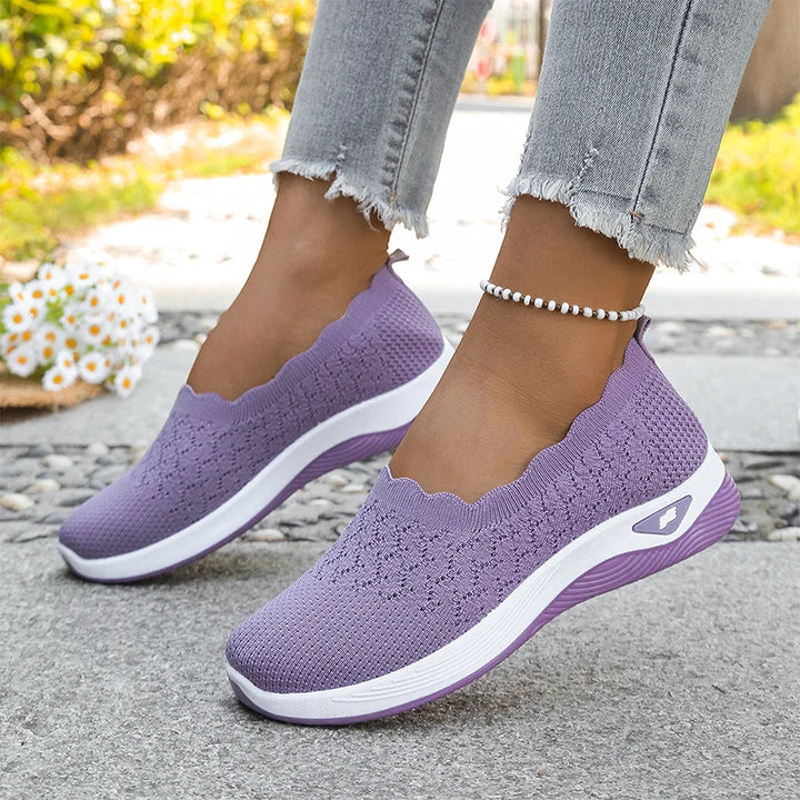Lilafarbene Strick-Slip-ons mit weißer Sohle an den Füßen auf Steinweg mit Blumen 19