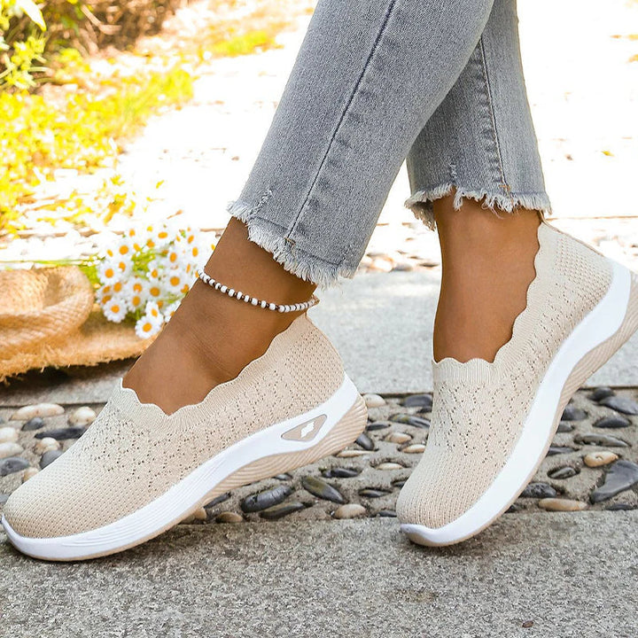 Beige Strick-Slip-ons mit Wellenrand an den Füßen auf Kieselsteinen, daneben Gänseblümchen und Strohhut 8