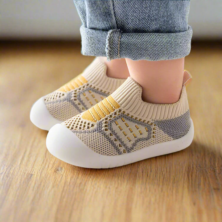 Seitenansicht beiger Strickschuhe mit gelben Streifen und grauen Elementen an Kinderfüßen auf Holzboden, Jeans umgekrempelt. 9