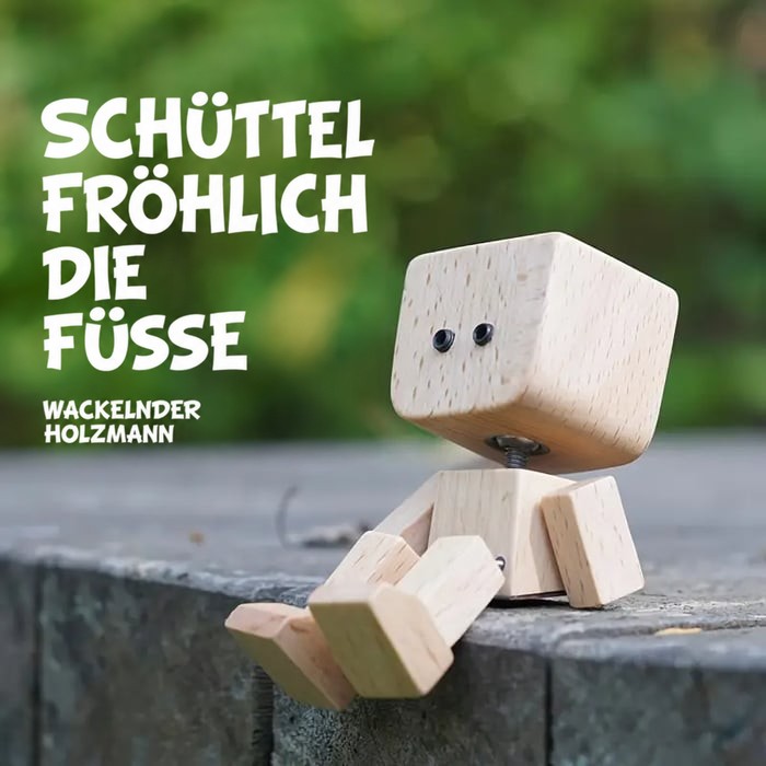 Holzfigur mit schlichten schwarzen Augen sitzt auf einer Steinkante vor grünem Hintergrund und lässt die Füße baumeln. 15