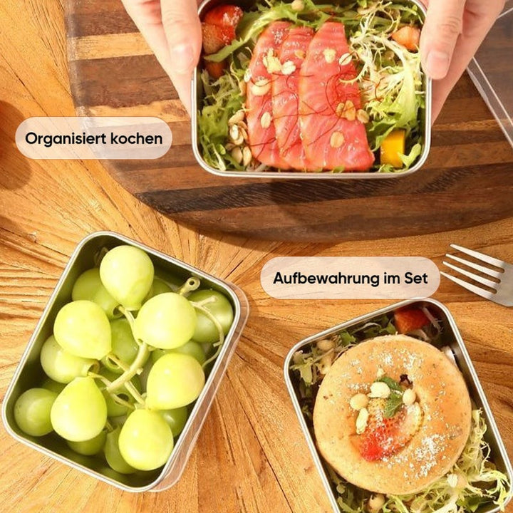 Draufsicht auf drei Edelstahlboxen auf einem Holztisch, gefüllt mit Lachs-Salat, grünen Trauben und einem belegten Gebäckstück, daneben eine Gabel. 8