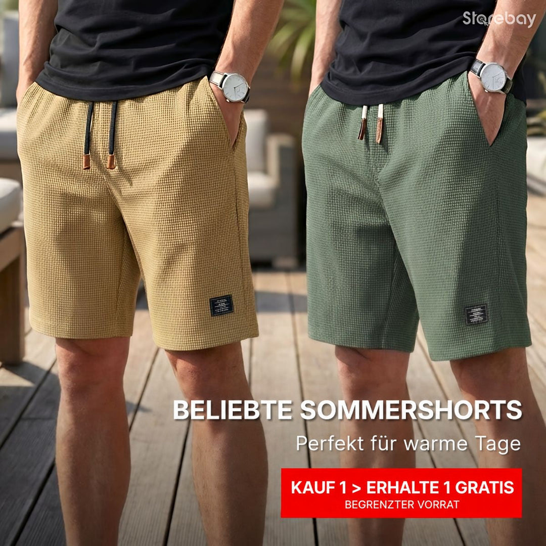 Zwei Männer in sandfarbener und grüner Waffelstruktur-Shorts mit Kordelzug, Hände in den Taschen, nebeneinander auf einer Holzterrasse. 1
