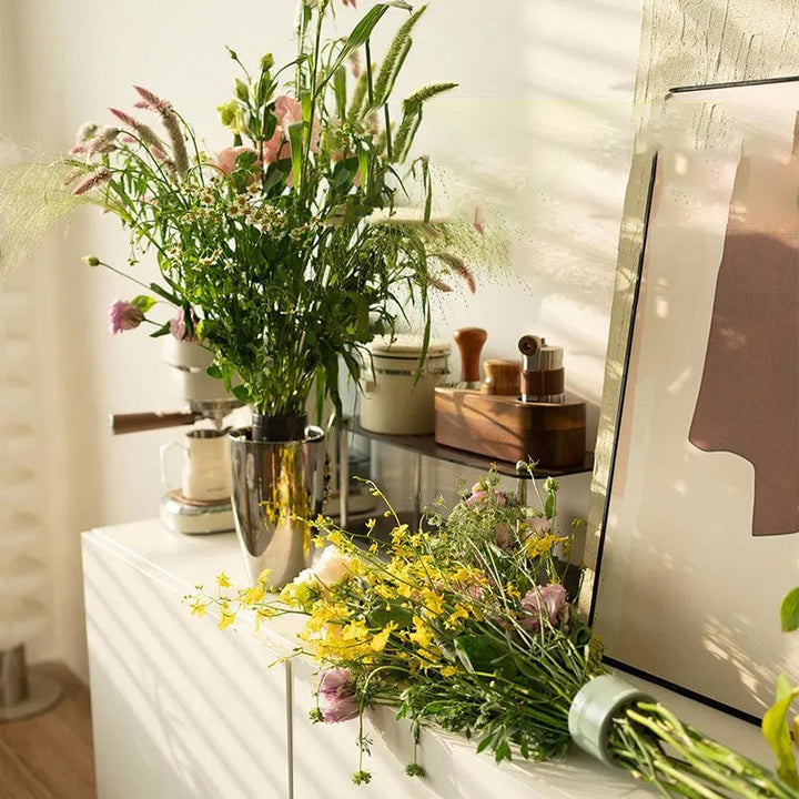 Wildblumenstrauß in einer Metallvase auf einer weißen Kommode, daneben liegt ein weiterer gebundener Strauß mit gelben Blüten und grünem Einsatz. 3