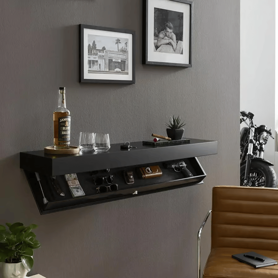 Schwarzes Wandregal an grauer Wand im Wohnraum, Fach aufgeklappt, oben Flasche, Gläser und Pflanze, daneben Bilderrahmen und Ledersessel. 4