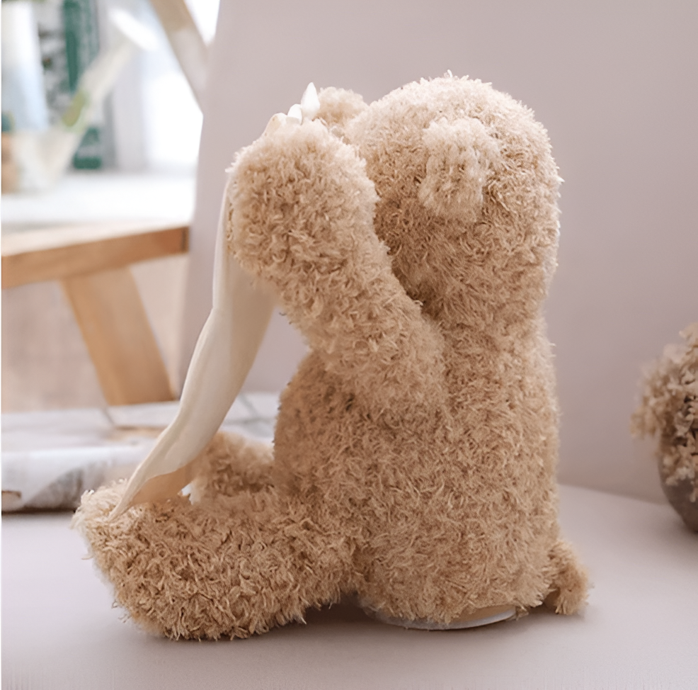 Seitenansicht von hinten: Der flauschige Bär sitzt auf einem Tisch und hebt einen Arm mit dem Tuch vors Gesicht. 3
