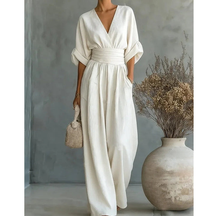 Cremeweißer Jumpsuit mit V-Ausschnitt und breitem Taillenband, Model mit Strohtasche und Trockenblumenvase im Bildausschnitt. 6