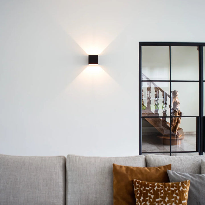 Schwarze Würfelleuchte an einer weißen Wand über einem grauen Sofa mit orangefarbenen Kissen, rechts eine Glastür mit Statue. 10