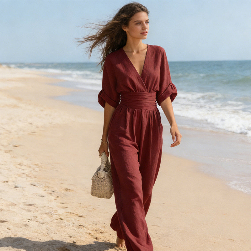 Model im weinroten Jumpsuit mit V-Ausschnitt beim Spaziergang am Sandstrand, Haare im Wind, Strohtasche in der Hand. 7