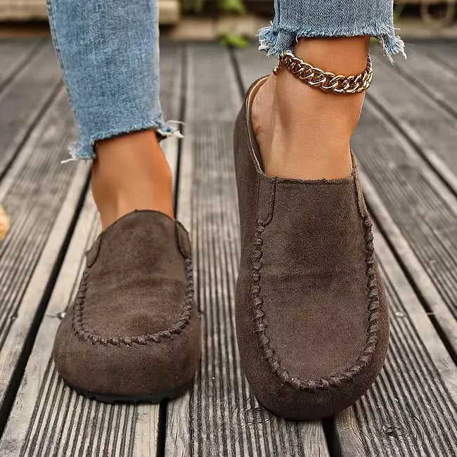 Frontansicht dunkelbrauner Slipper an den Füßen einer Frau in Jeans mit Fußkettchen, die flache Sohle und die Ziernaht erkennbar. 4