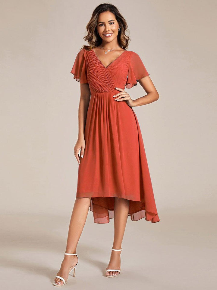 Model in terrakottafarbenem Chiffonkleid mit Wickeloptik, Flatterärmeln und Hoch-Tief-Saum, Hand an der Hüfte, weiße Sandaletten. 92
