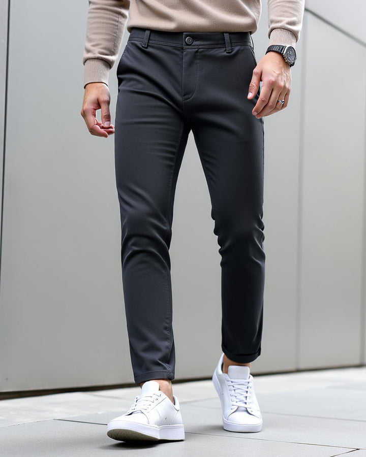 Dunkelgraue Chino mit umgeschlagenem Saum, getragen mit beigem Strickpulli, Armbanduhr und weißen Sneakern vor grauer Wand. 9