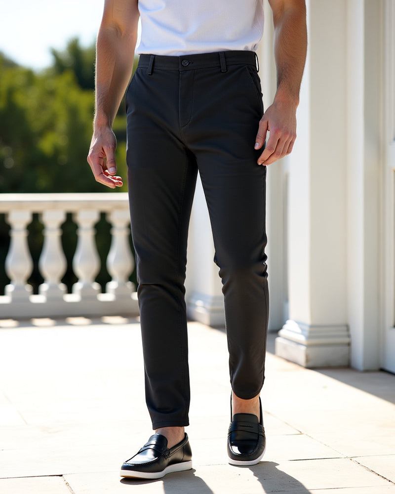 Dunkelgraue Chino auf sonniger Terrasse vor weißer Balustrade, kombiniert mit weißem Shirt und schwarzen Lederslippern. 7