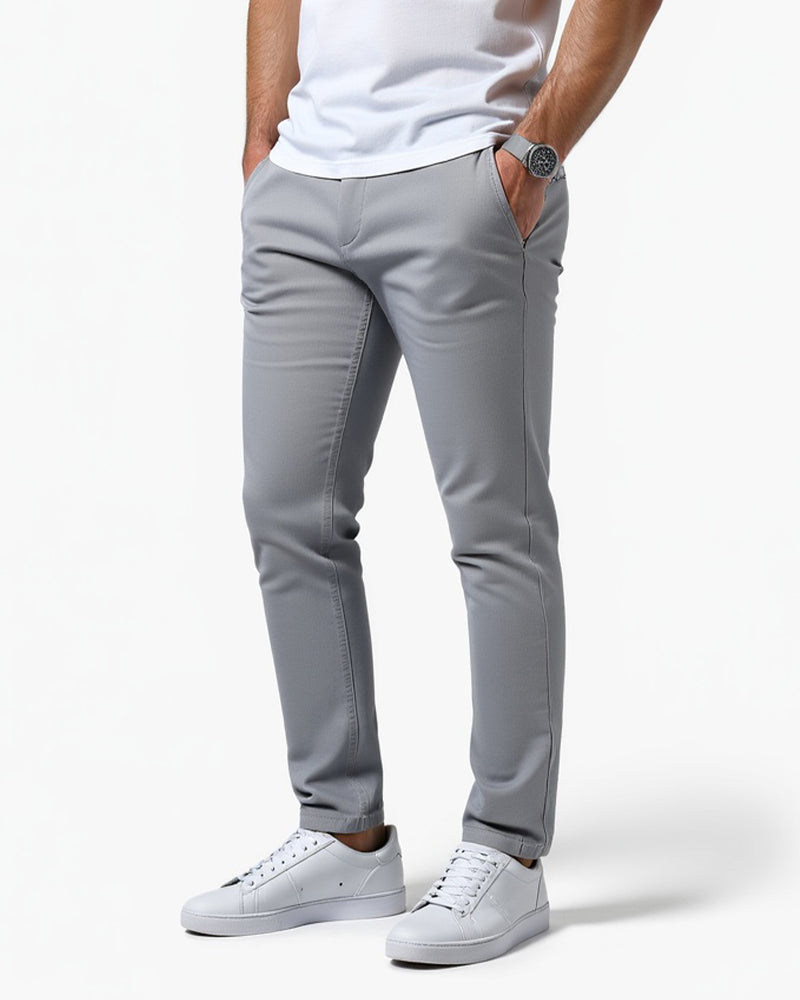 Hellgraue Chino in Seitenansicht mit weißem T-Shirt und weißen Sneakern, eine Hand in der Hosentasche, Armbanduhr sichtbar. 11