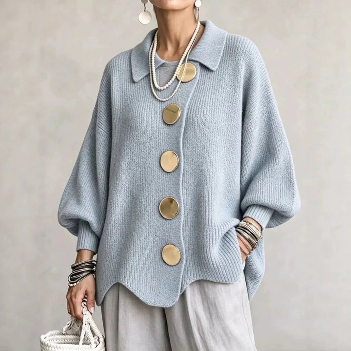 Frau trägt die Strickjacke in hellem Graublau mit goldfarbenen Knöpfen, grauer Hose und Armreifen, Frontansicht. 5