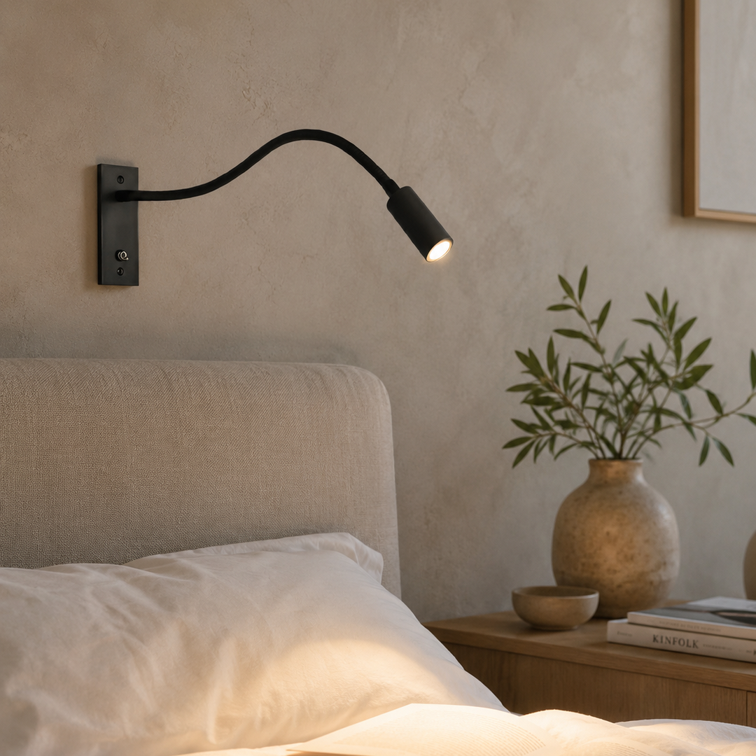 Eingeschaltete schwarze Wandlampe über dem Bett in warmem Abendlicht, davor ein aufgeschlagenes Buch, an der Wand eine Vase mit Zweigen. 3