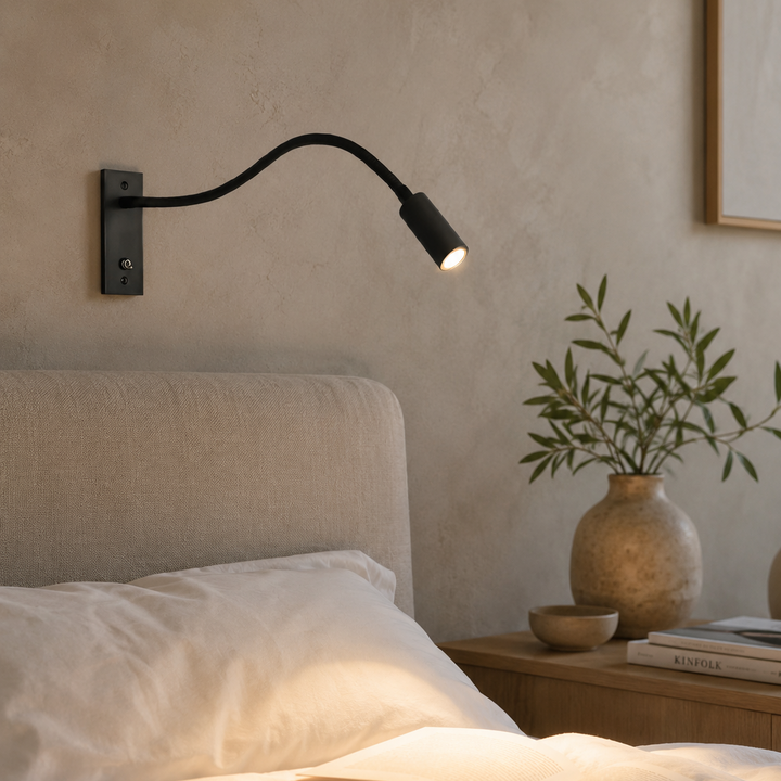 Eingeschaltete schwarze Wandlampe über dem Bett in warmem Abendlicht, davor ein aufgeschlagenes Buch, an der Wand eine Vase mit Zweigen. 3