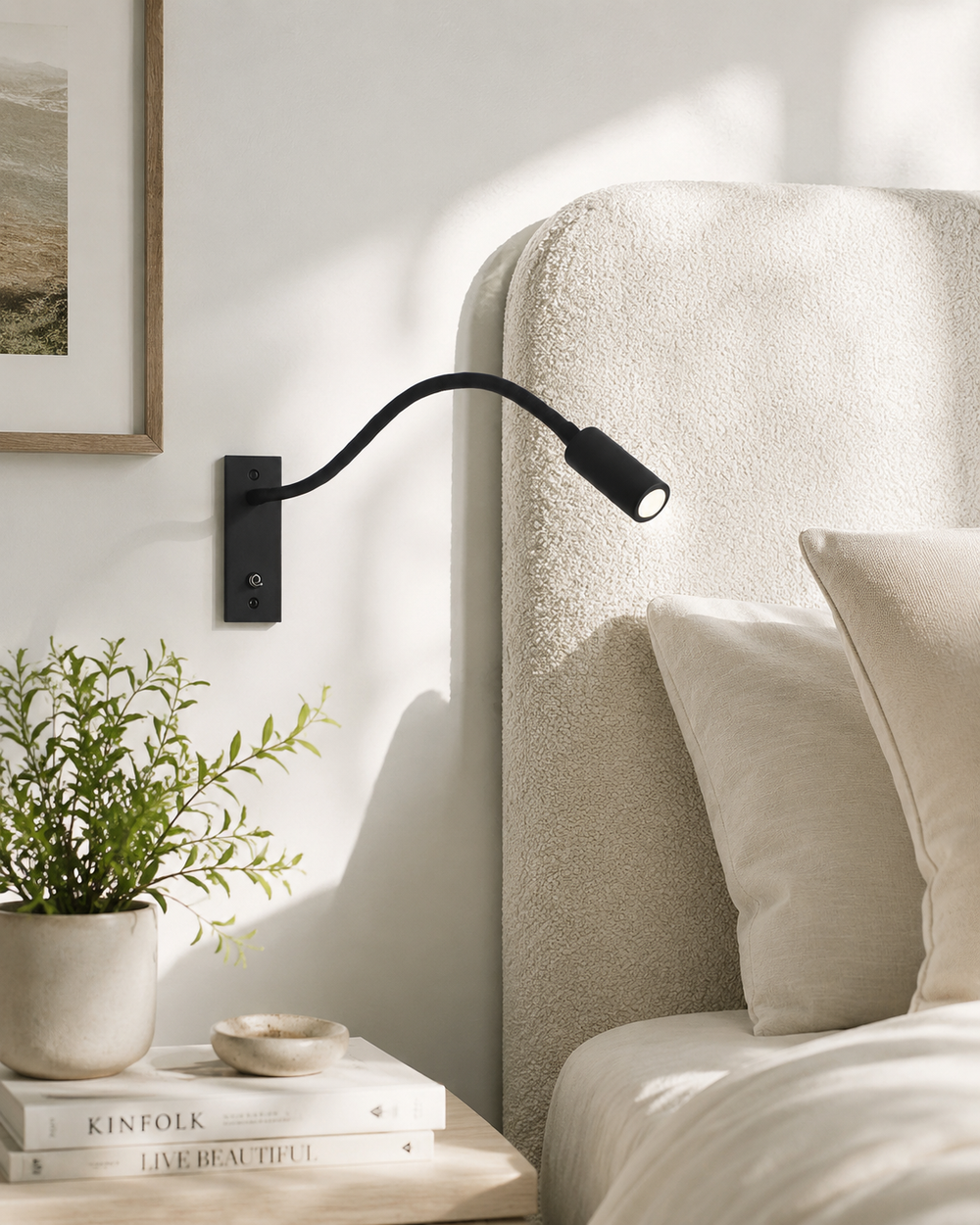 Schwarze Leselampe an heller Wand neben einem Bett mit cremefarbenem Bouclé-Kopfteil, daneben Nachttisch mit Pflanze und Büchern. 2