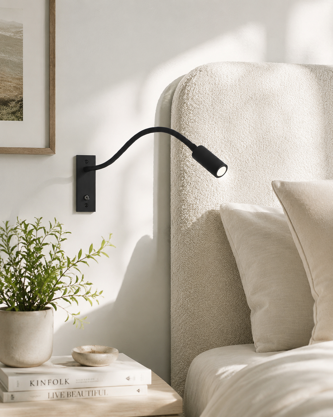 Schwarze Leselampe an heller Wand neben einem Bett mit cremefarbenem Bouclé-Kopfteil, daneben Nachttisch mit Pflanze und Büchern. 2