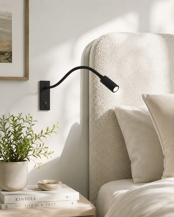 Schwarze Leselampe an heller Wand neben einem Bett mit cremefarbenem Bouclé-Kopfteil, daneben Nachttisch mit Pflanze und Büchern. 2