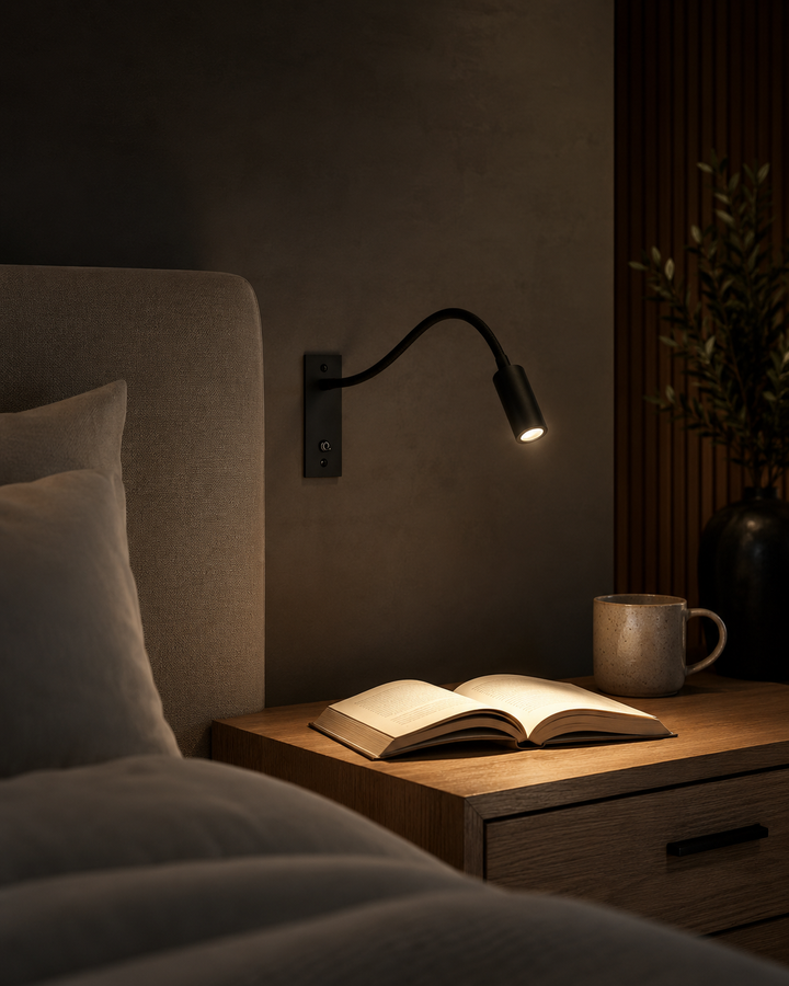 Schwarze Wandlampe in abgedunkeltem Schlafzimmer beleuchtet ein aufgeschlagenes Buch auf dem Holznachttisch, daneben eine Tasse. 6