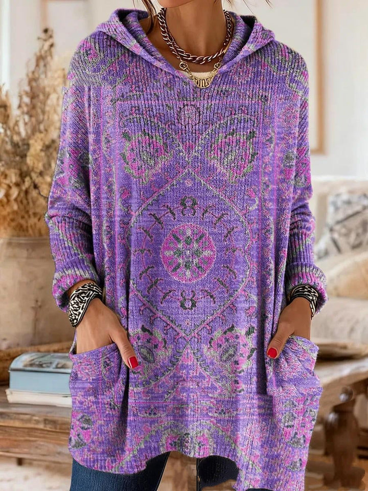 Getragene Kapuzentunika in Violett mit rosa und grünem Ornamentmuster, Hände in den großen Vordertaschen, Nahaufnahme von Oberkörper und Saum. 2