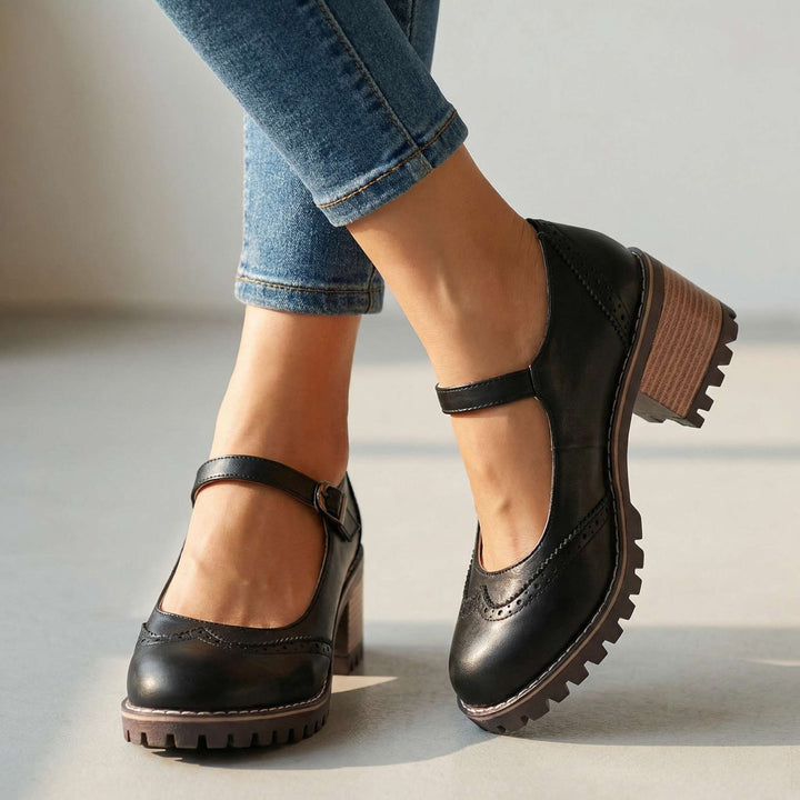 Schwarze Mary Janes mit Lochmuster, Schnallenriemen, Blockabsatz und Profilsohle, getragen zu Jeans vor hellem Studiohintergrund. 1