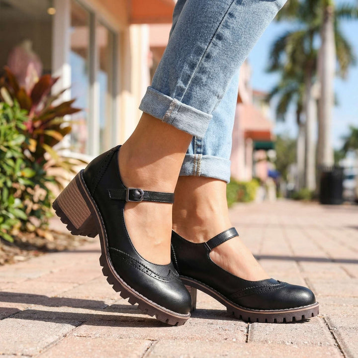Schwarze Mary Janes mit verstellbarem Riemen, getragen zu hellblauer Jeans auf einem gepflasterten Gehweg mit Palmen im Hintergrund. 4
