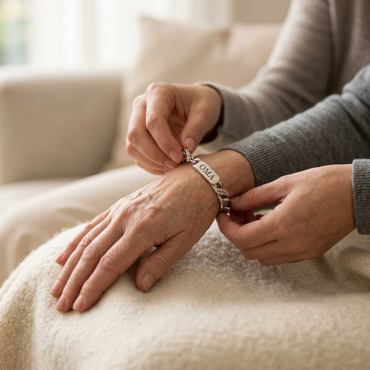 Eine Person legt einer älteren Frau das silberfarbene Armband mit graviertem Mittelstück um das Handgelenk, auf einem Sofa mit Decke. 3