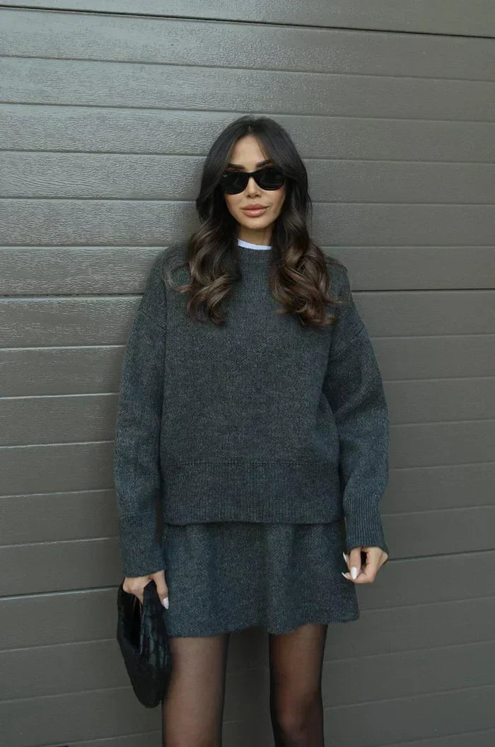 Model in anthrazitgrauem Strick-Set, Pullover mit breitem Saum und kurzer Rock, dazu Strumpfhose und schwarze Clutch. 11