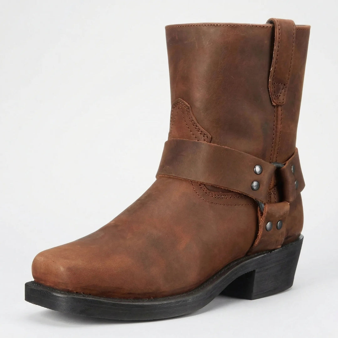 Brauner Lederstiefel mit genietetem Zierriemen, eckiger Spitze und schwarzem Absatz, schräg von vorn vor hellem Hintergrund. 2