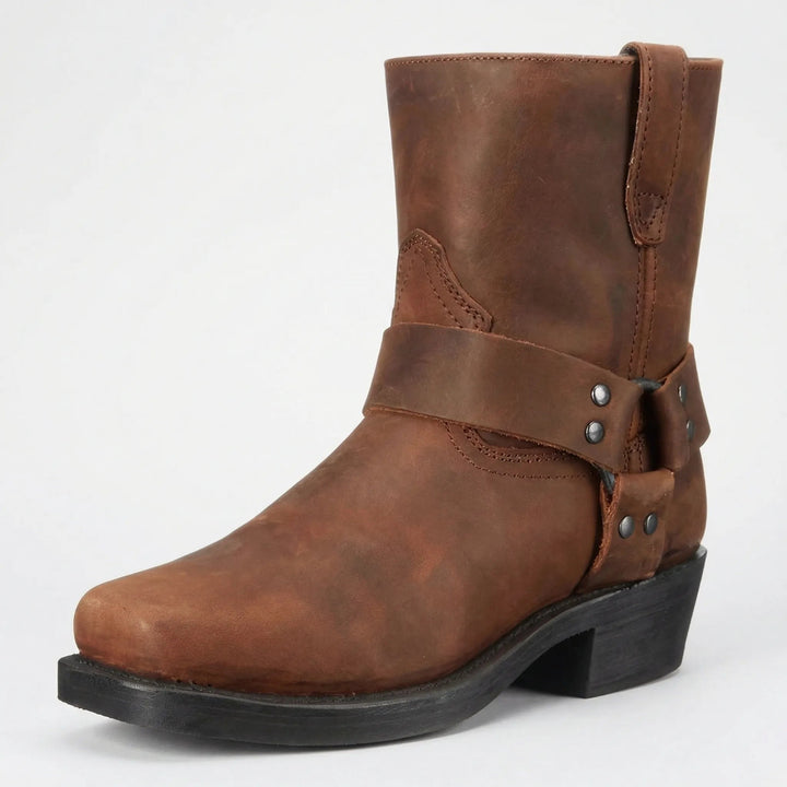 Brauner Lederstiefel mit genietetem Zierriemen, eckiger Spitze und schwarzem Absatz, schräg von vorn vor hellem Hintergrund. 2