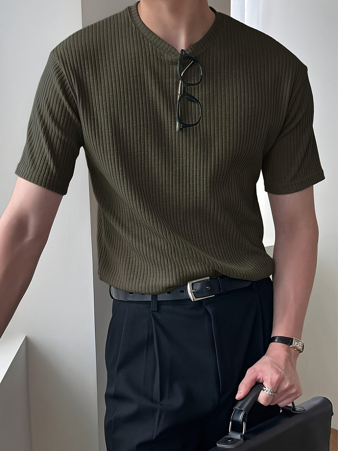 Mann in olivgrünem, geripptem T-Shirt mit schwarzer Hose und Brille, hält schwarze Aktentasche.