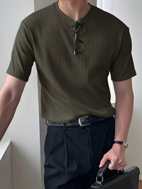 Mann in olivgrünem, geripptem T-Shirt mit schwarzer Hose und Brille, hält schwarze Aktentasche.
