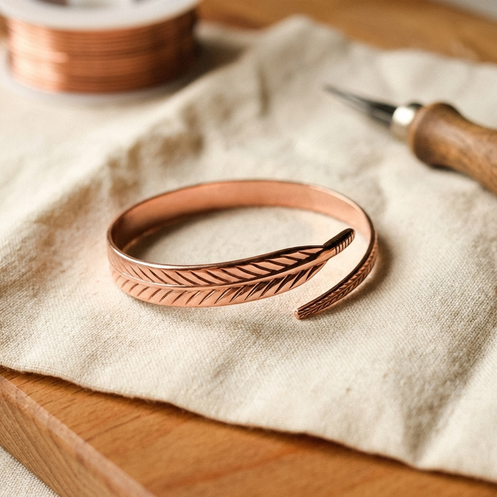 Offenes Kupfer-Armband mit eingraviertem Federmuster auf einem Leinentuch, daneben eine Kupferdrahtspule und ein Graviergriff mit Holzgriff. 1