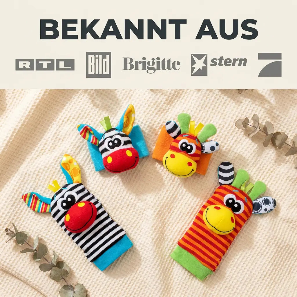 Zwei Handgelenkrasseln und zwei Rasselsocken mit Zebra- und Giraffengesicht liegen auf einer cremefarbenen Waffelstrick-Decke, daneben Eukalyptuszweige. 7