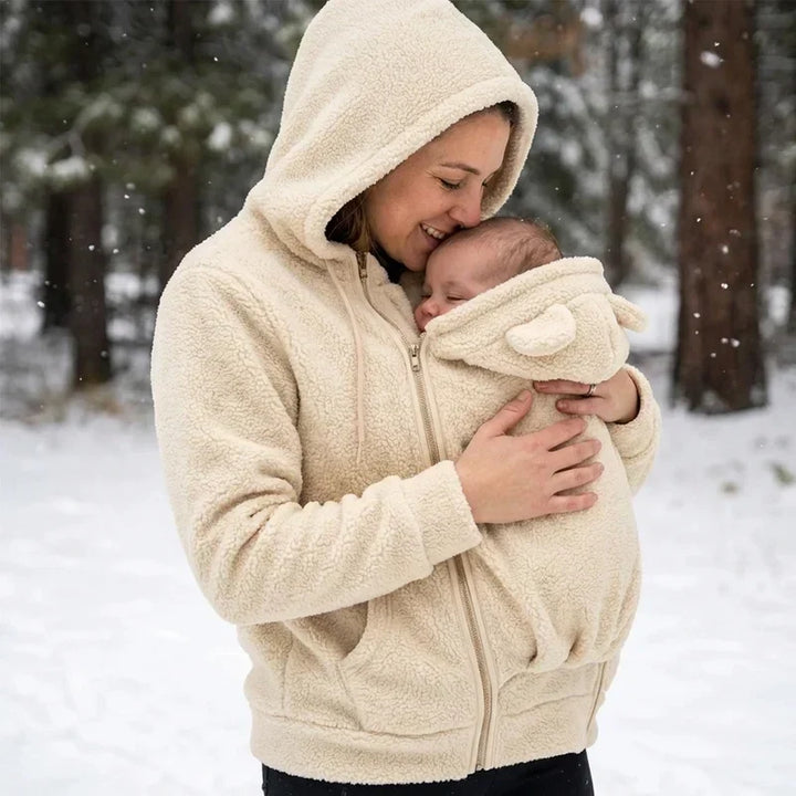 Frau im beigen Sherpa-Hoodie küsst im verschneiten Wald ihr Baby auf die Stirn, das im Trageeinsatz mit Öhrchen geborgen liegt. 8
