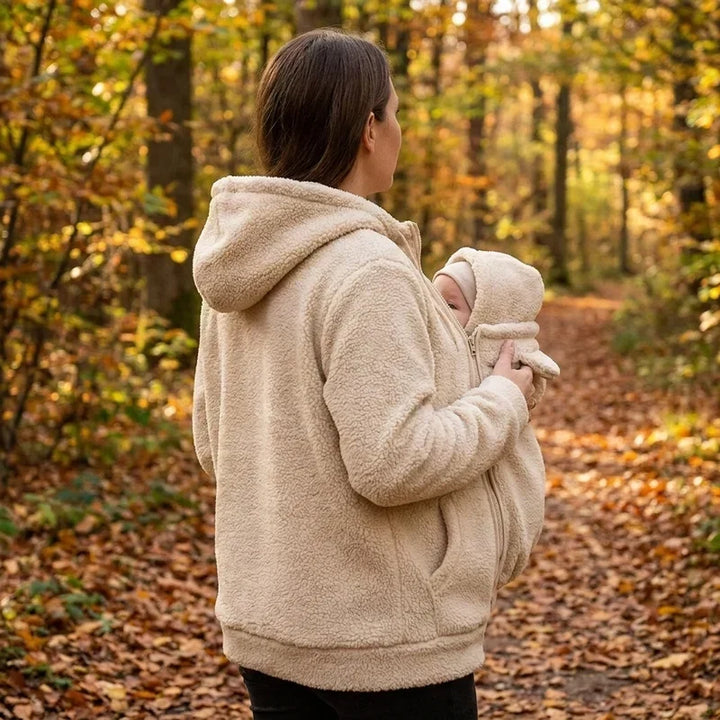 Rückansicht einer Frau im beigen Sherpa-Hoodie im Herbstwald, seitlich ist das Baby im Trageeinsatz mit Kapuze zu sehen. 6