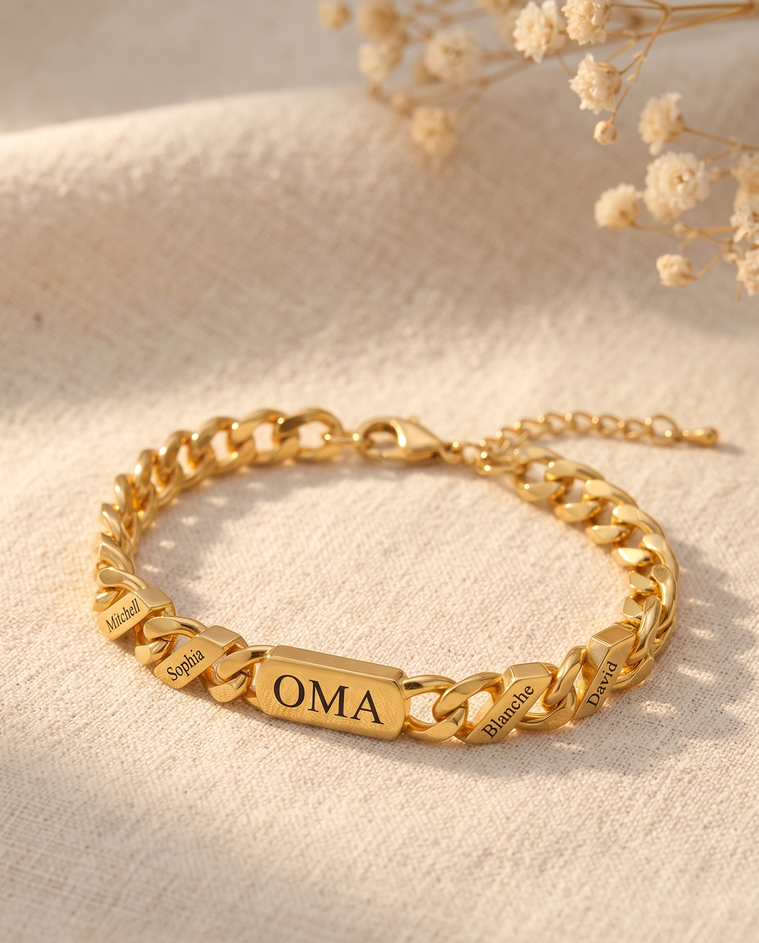 Goldfarbenes Gliederarmband mit mehreren gravierten Namensgliedern liegt auf hellem Leinenstoff, daneben getrocknete Blüten und die Verlängerungskette. 4
