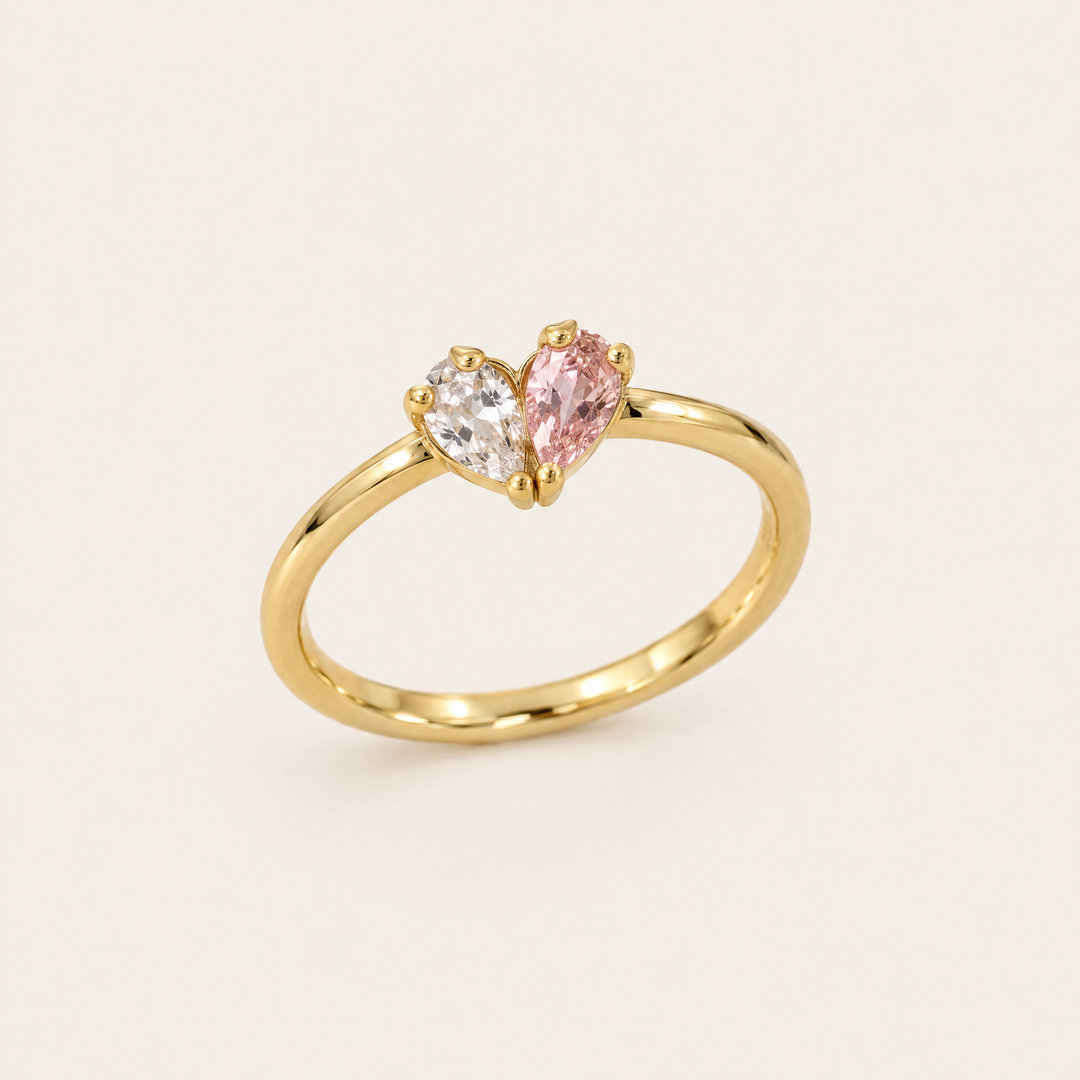 Goldfarbener Ring in Schrägansicht, dessen zwei tropfenförmige Steine, ein klarer und ein rosafarbener, zusammen ein Herz bilden. 1