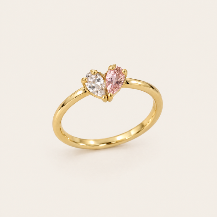 Goldfarbener Ring in Schrägansicht, dessen zwei tropfenförmige Steine, ein klarer und ein rosafarbener, zusammen ein Herz bilden. 1