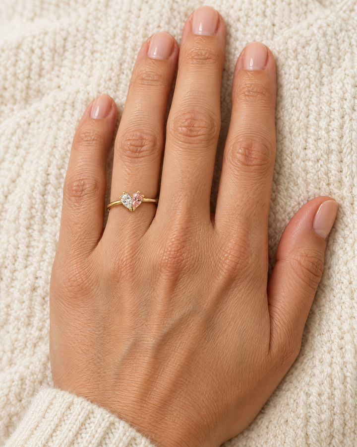 Der Ring mit klarem und rosafarbenem Stein am Finger einer Hand, die auf einem cremefarbenen Strickstoff ruht. 2