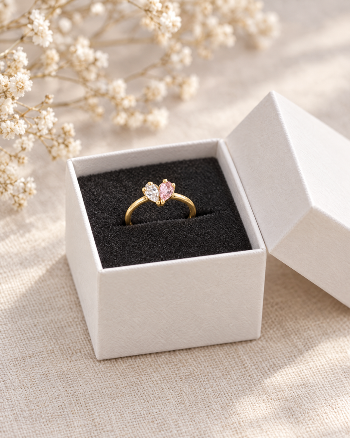 Der goldfarbene Ring liegt in einer offenen weißen Schmuckschachtel mit schwarzem Einsatz, daneben getrocknete Blüten. 3