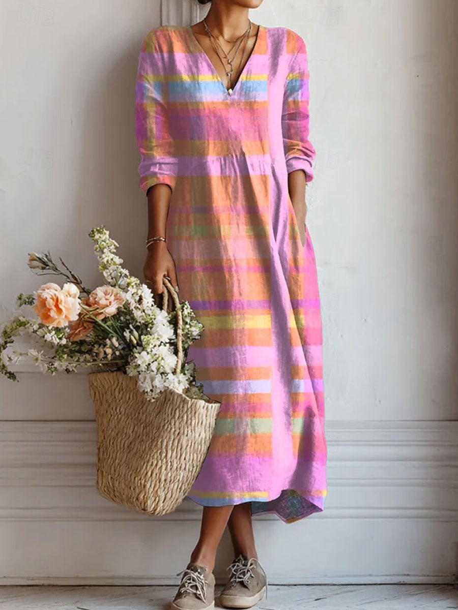 Frau trägt ein langärmliges Midikleid mit V-Ausschnitt und buntem Karomuster in Rosa und Orange, eine Hand in der Tasche, dazu Strohkorb mit Blumen. 1