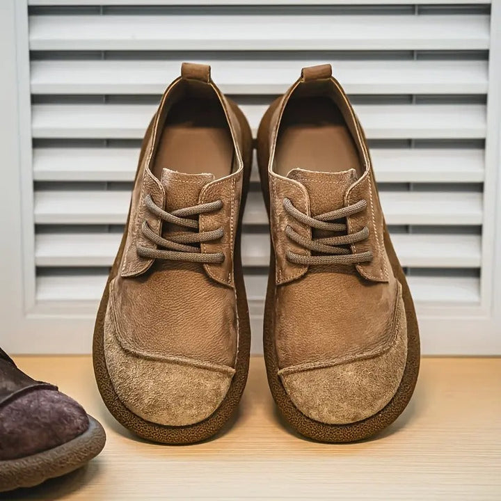 Paar hellbraune Schnürschuhe aus Leder mit Wildleder-Kappe und breiter Gummisohle, frontal vor einer Jalousie auf einem Holzboden aufgestellt. 1