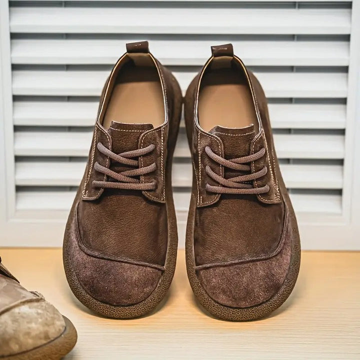 Dunkelbraune Schnürschuhe mit Wildleder-Kappe und Ziernähten, frontal vor einer Jalousie auf einem Holzboden aufgestellt. 10